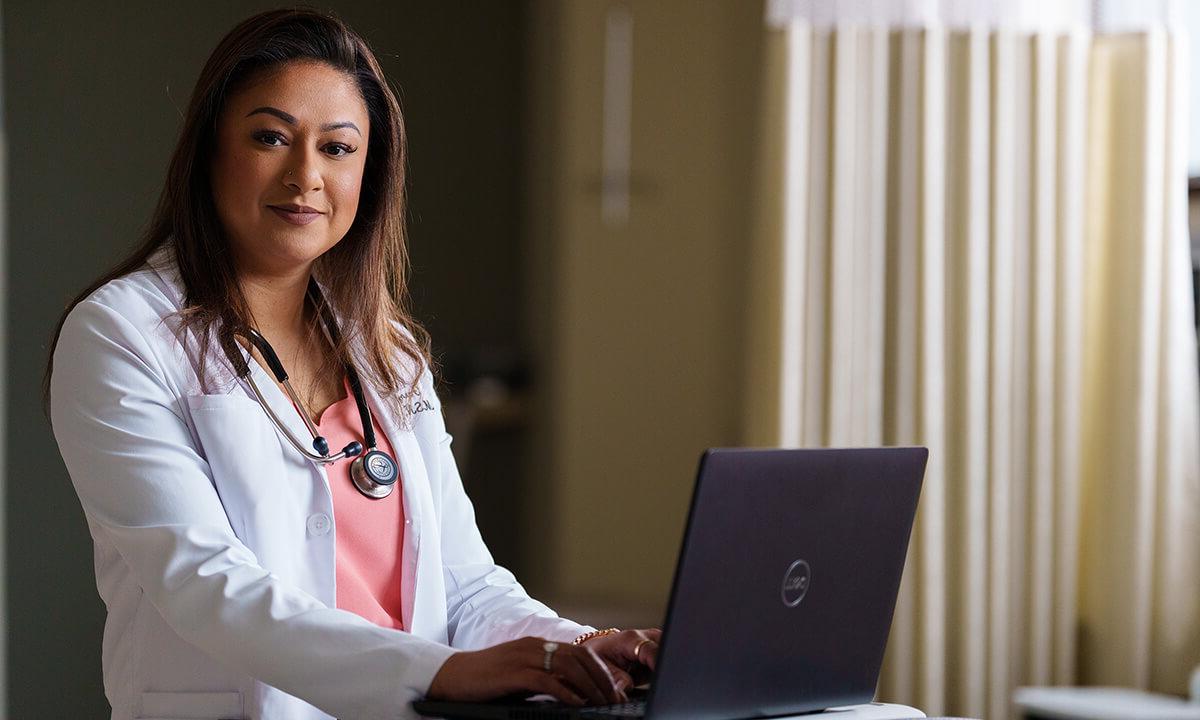 A woman in a medical coat sits before a laptop.